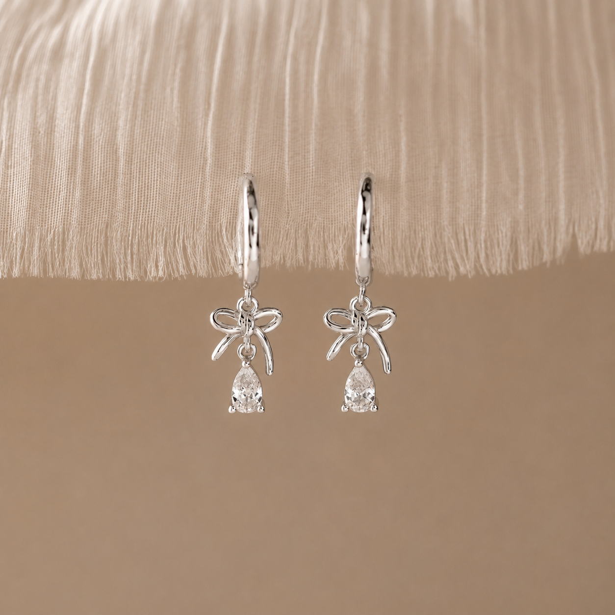 Silver earrings with bow designs and teardrop-shaped stones on a beige fabric background
